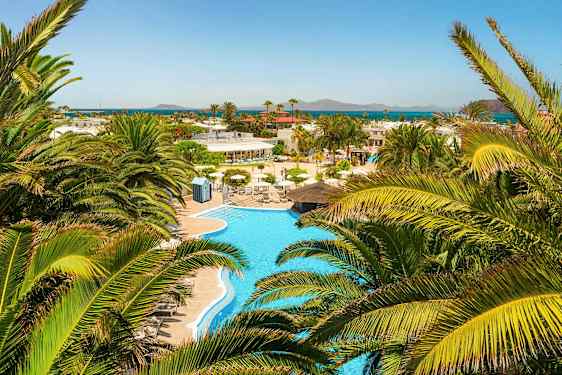 Et udsyn over en swimmingpool omgivet af palmer med udsigt til havet i det fjerne. (AI-genereret tekst)