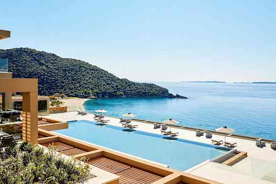 Udsigt over en pool med solsenge og parasoller, en kystlinje og hav i baggrunden. (AI-genereret tekst)