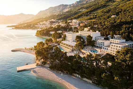 Luftfoto af hotel ved kysten og strand, omgivet af træer og med bjerge i baggrunden. (AI-genereret tekst)