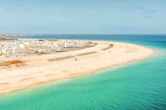 Luftfoto af en hvid sandstrand med klart turkisblåt vand og hvide bygninger i baggrunden. (AI-genereret tekst)