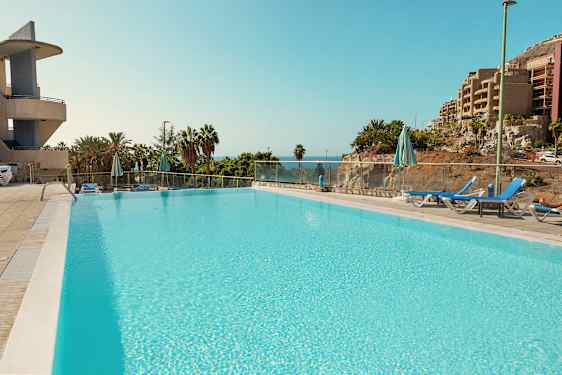 En stor pool omgivet af liggestole og parasoller, med udsigt til havet og frodig vegetation. (AI-genereret tekst)