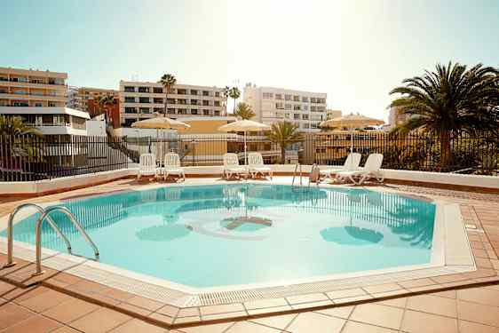 Udendørs pool med liggestole og parasoller, omgivet af bygninger og palmer. (AI-genereret tekst)