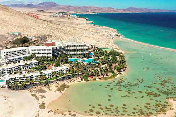 Hotelkompleks ved en strand med turkis vand og omgivelser af ørkenlandskab. (AI-genereret tekst)