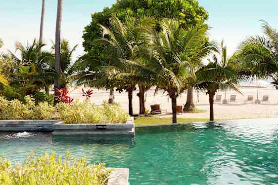 Poolområde med udsigt til en sandstrand og omkringliggende palmer. (AI-genereret tekst)