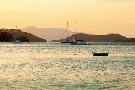 Billede af en stille bugt ved solnedgang med sejlbåde og en grøn båd i forgrunden. (AI-genereret tekst)