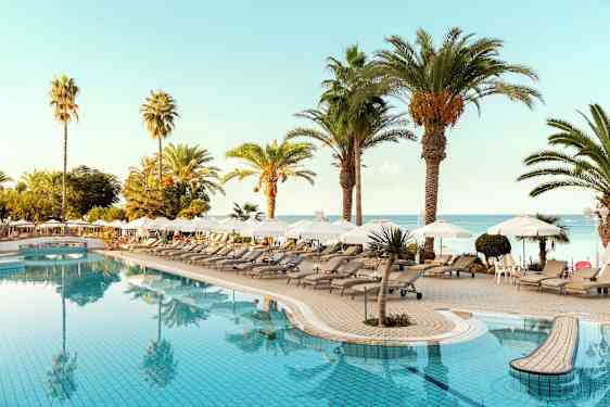 Poolområde med liggestole, parasoller og palmer tæt på havet. (AI-genereret tekst)