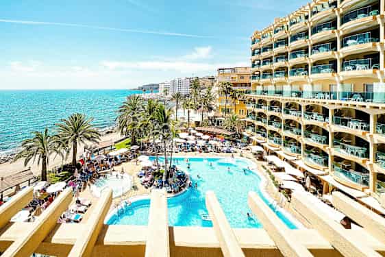 En solrig hotelhave med swimmingpools, palmer og udsigt over havet. (AI-genereret tekst)