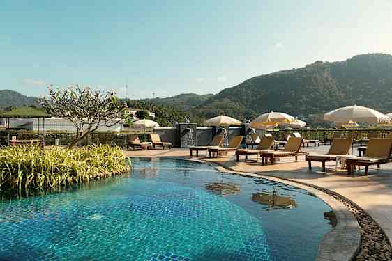 Udendørs pool med liggestole og parasoller, omgivet af grønne områder og bjerge i baggrunden. (AI-genereret tekst)