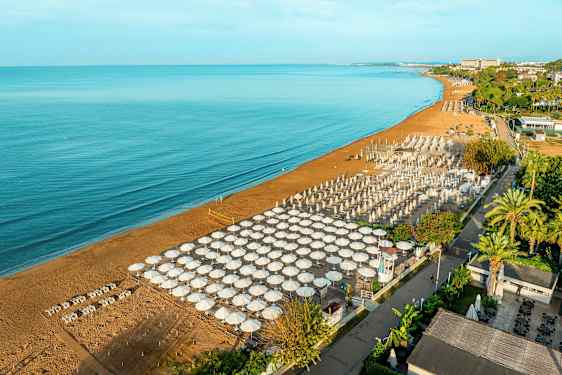 Billedet viser en sandstrand ved en hav, med mange parasoller og liggestole opstillet i en organiseret formation. I baggrunden ses grønne træer og bygninger. (AI-genereret tekst)