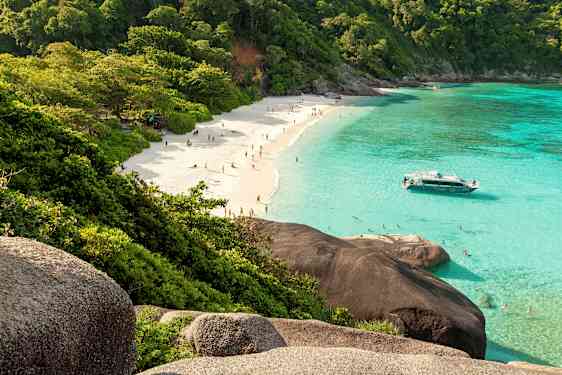 En sandstrand med mennesker, omgivet af frodige træer og klart turkis vand. En båd ligger tæt på stranden. (AI-genereret tekst)