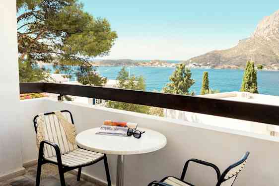 En balkon med stole og bord, hvorpå der ligger en bog og solbriller. Udsigt over havet og en bjergagtig baggrund. (AI-genereret tekst)