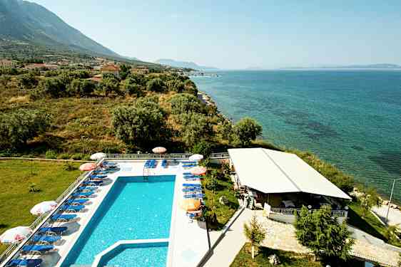 Udendørs pool med solvogne ved siden af havet, omgivet af grønt og bygninger. (AI-genereret tekst)