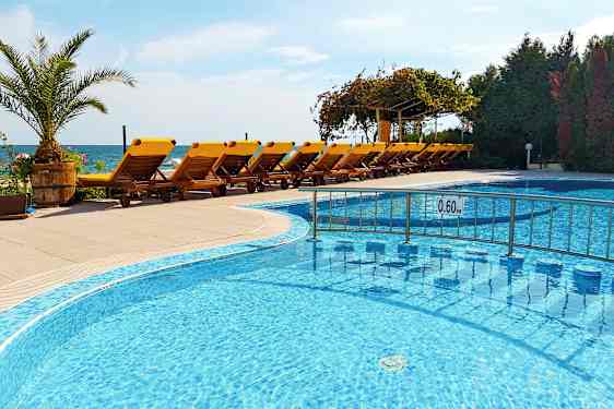 En swimmingpool med lavt vand foran en række gule liggestole på en terrasse, med havet i baggrunden. (AI-genereret tekst)