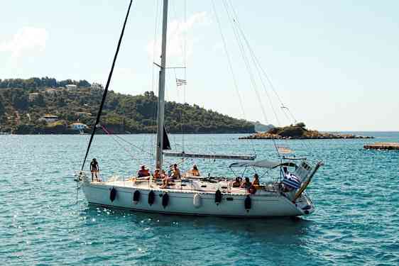 En sejlbåd med flere personer om bord sejler tæt på en kystlinje med grønne bakker og nogle huse. (AI-genereret tekst)