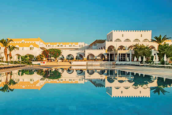 En hvid bygning med gult tag ved en pool med solsenge og parasoller. (AI-genereret tekst)