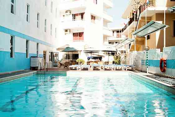 Udendørs pool med liggestole og parasoller, omgivet af bygninger. (AI-genereret tekst)