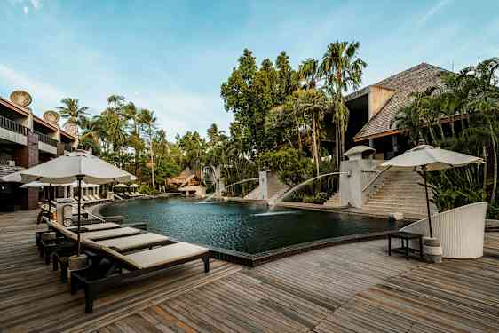 En udendørs pool omgivet af trædæk og liggestole med parasoller. Der er frodig beplantning og bygninger af træ og sten i baggrunden. (AI-genereret tekst)