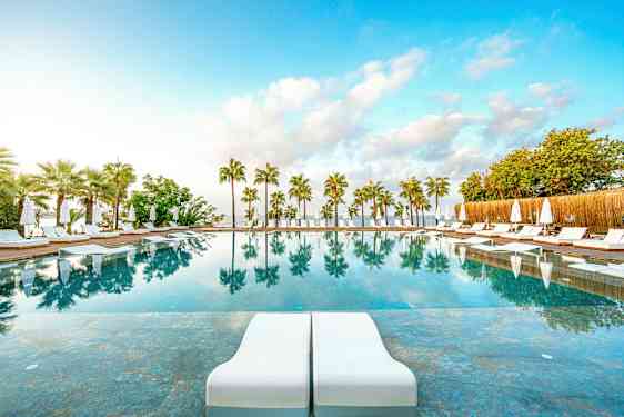 En udendørs pool med solsenge og parasoller, omgivet af palmer, og med spejlinger i vandet. (AI-genereret tekst)