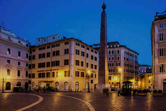 En plads med bygninger omkring og en obelisk i midten, oplyst om natten. (AI-genereret tekst)