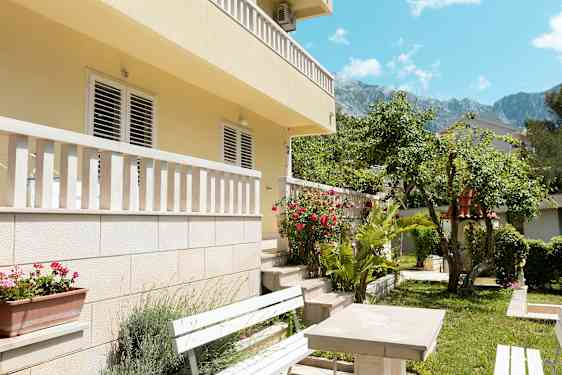 Billede af en husfacade med altan og have i forgrunden, inklusive bænke og blomster. (AI-genereret tekst)