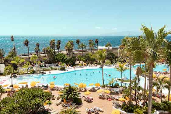 Poolområde med liggestole, parasoller og mennesker, med havet i baggrunden. (AI-genereret tekst)