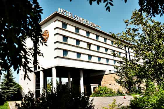 Bygning med flere etager, mærket med teksten 'Hotel Sympozjum', omgivet af træer og vegetation. (AI-genereret tekst)