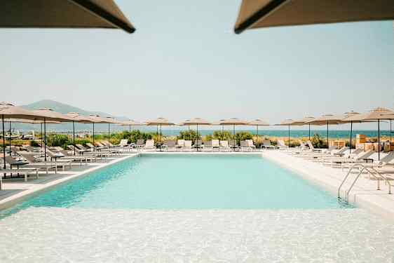 En udendørs pool omgivet af liggestole og parasoller, med udsigt over landskab og hav. (AI-genereret tekst)