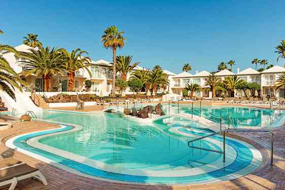 En hotelpool omgivet af palmer, liggestole og hvide bygninger. (AI-genereret tekst)