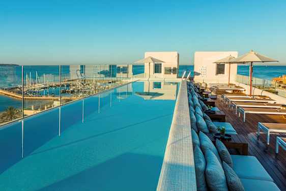 Udsigt over en infinitypool med liggestole og parasoller, omgivet af glasrækværk og en marina i baggrunden. (AI-genereret tekst)