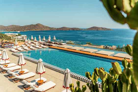 En pool med liggestole og parasoller ved en kystlinje med udsigt over et blåt hav og bjerge. (AI-genereret tekst)