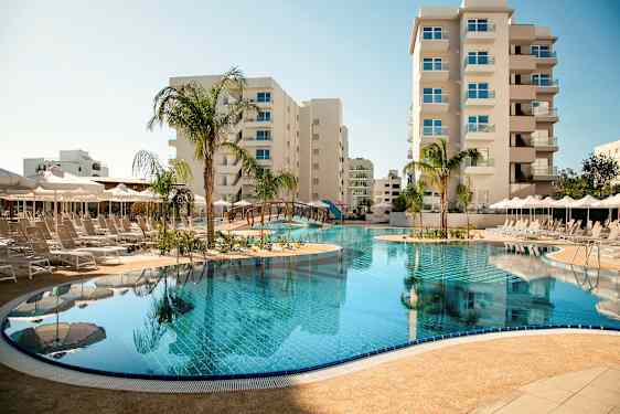 En udendørs pool omgivet af liggestole og parasoller med høje bygninger i baggrunden. (AI-genereret tekst)