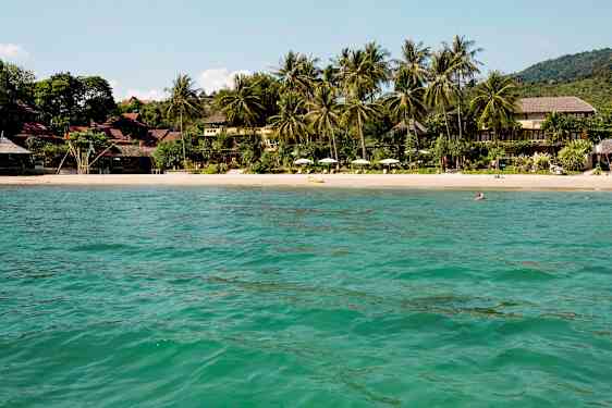 Udsigt over en sandstrand omgivet af tropiske træer og bygninger, set fra vandet. (AI-genereret tekst)