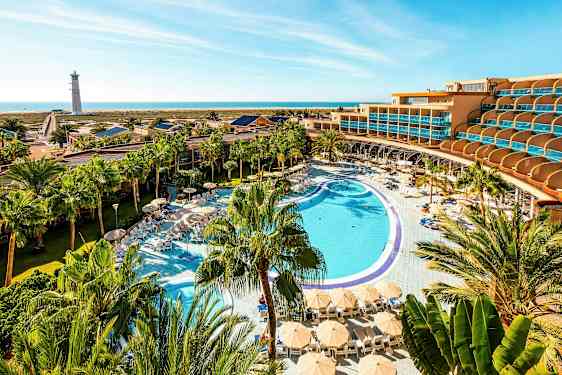 En hotelkompleks med en stor pool, liggestole og parasoller omgivet af palmer, med havet og en fyr i baggrunden. (AI-genereret tekst)