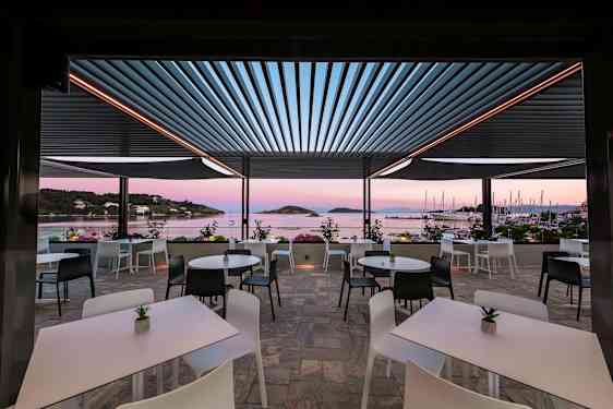 En udendørs terrasse med borde og stole med udsigt over havet og sejlbåde. (AI-genereret tekst)