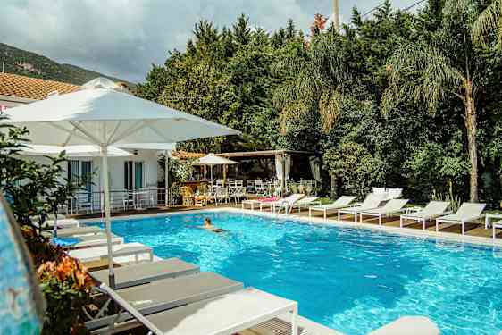 En udendørs pool omgivet af hvide liggestole og parasoller nær en bygning og grønne områder. (AI-genereret tekst)