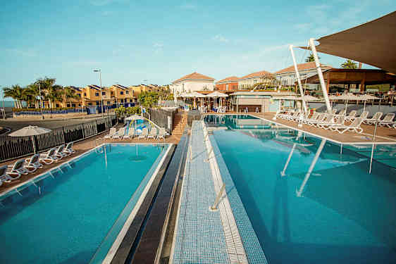 Et billede af et solrigt feriested med to udendørspools omgivet af liggestole med bygninger i baggrunden. (AI-genereret tekst)