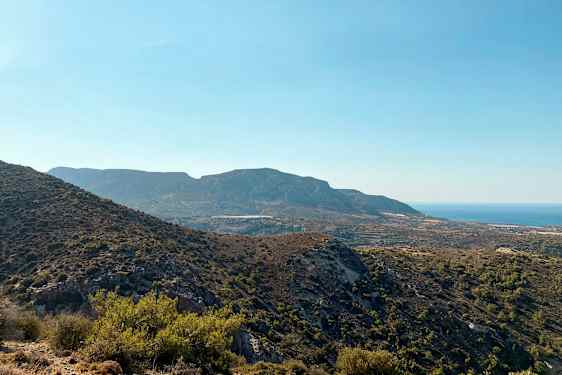 En udsigt over et bjergrigt landskab med varieret vegetation og havet i baggrunden under en klar blå himmel. (AI-genereret tekst)