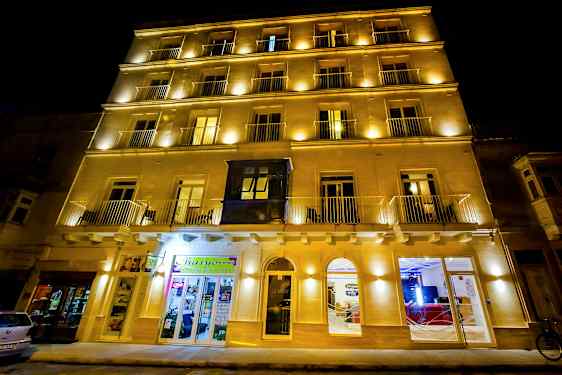 En bygning fotograferet om natten med oplyste facader og flere vinduer. Nogle butikker og indgange er synlige. (AI-genereret tekst)