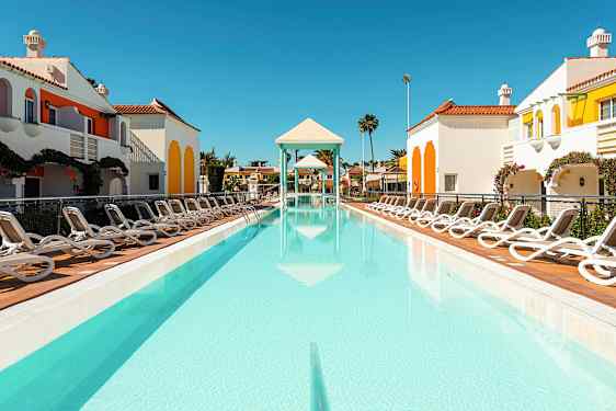 En udendørs pool omgivet af liggestole og farverige bygninger med en klar himmel. (AI-genereret tekst)