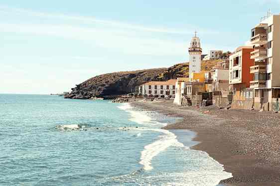 Billede af en kyst med sorte sandstrande, et fyrtårn og bygninger ved vandkanten. (AI-genereret tekst)