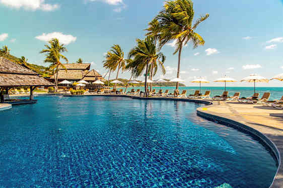 Billede af en udendørs pool omgivet af liggestole og palmer, med havet i baggrunden. (AI-genereret tekst)