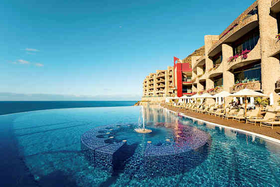 En infinitypool ved strandkanten med solsenge og hotelbygninger i baggrunden. (AI-genereret tekst)