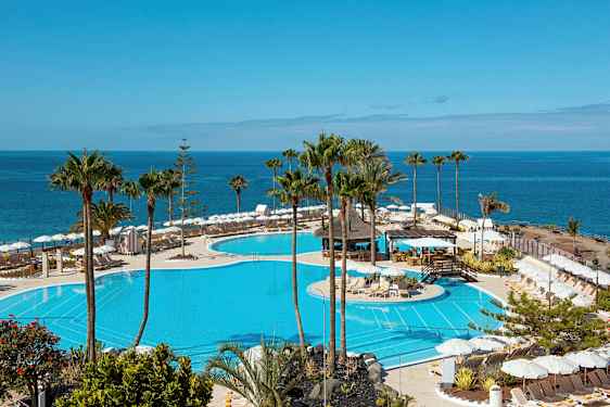 Udsigt over en stor pool med liggestole og parasoller omkring samt havet i baggrunden. (AI-genereret tekst)