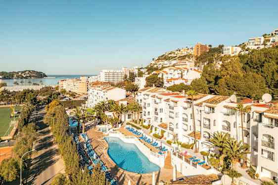 Billede af hotelområde med pool og liggestole, omgivelser med træer og bygninger samt udsigt mod havet. (AI-genereret tekst)