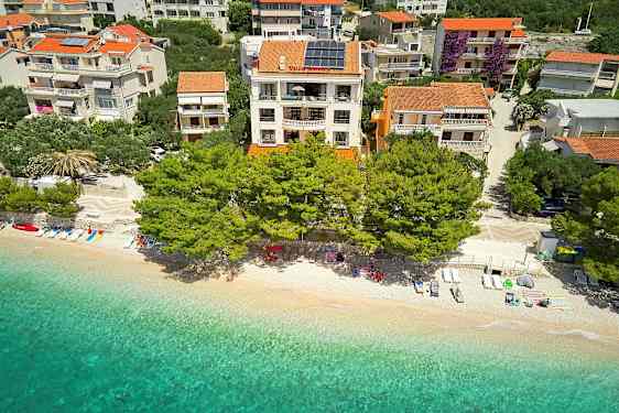 Luftfoto af en strand med liggestole, parasoller og klart vand foran bygninger med terrakottatag omgivet af træer. (AI-genereret tekst)