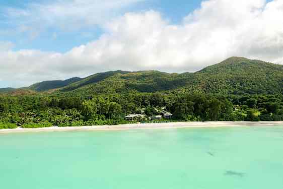 En strand med hvidt sand foran en skovklædt bjergkæde og turkisfarvet vand. (AI-genereret tekst)