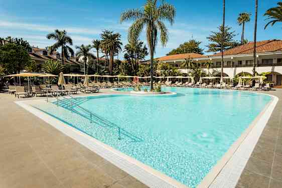 En udendørs pool omgivet af liggestole og parasoller med palmer i baggrunden. (AI-genereret tekst)