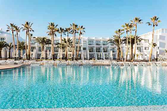 Hotellets poolområde med klart blåt vand, omgivet af liggestole, parasoller og palmer. I baggrunden ses en hvid bygning med altaner. (AI-genereret tekst)