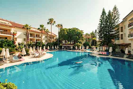 Stor udendørs swimmingpool omgivet af liggestole og parasoller, med grønne træer og hotelbygninger i baggrunden. (AI-genereret tekst)