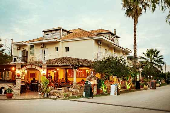Restaurant med udendørs veranda i stueetagen af en lys bygning, omgivet af græs og palmer. (AI-genereret tekst)